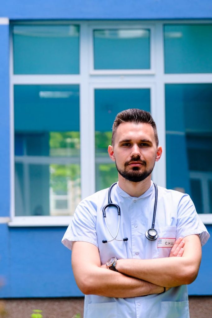 Bearded doctor with stethoscope standing confidently outside a blue hospital building.